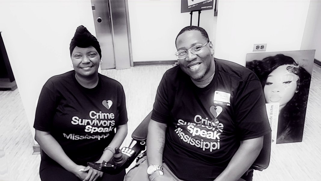 Two participants seated indoors wearing Crime Survivors Speak Mississippi shirts. The second person is using a motorized scooter. A large portrait of a violence victim is to the right of them.
