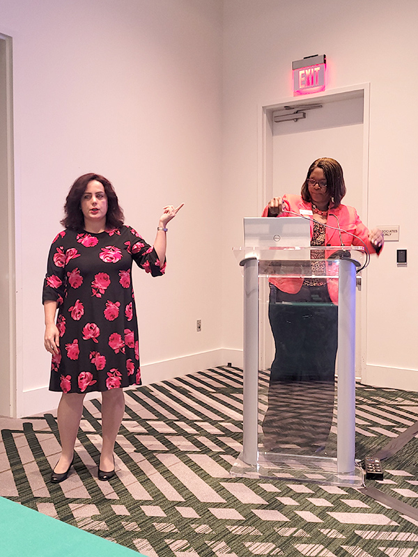 Conference presentation with a speaker at a podium and a staff member standing nearby, in a meeting room with patterned carpeting and presentation equipment.