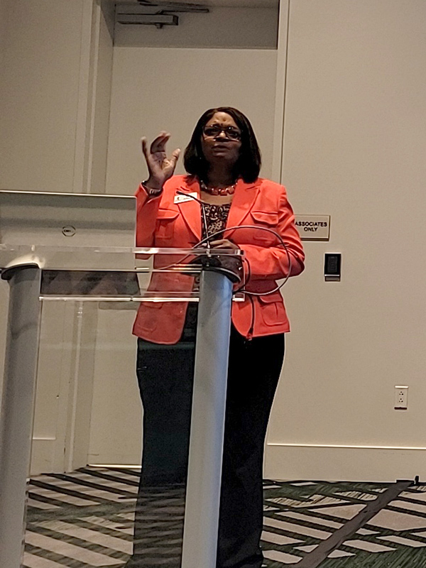 Conference speaker standing at a clear podium, gesturing while delivering a presentation in a meeting room.