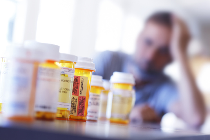 A large group of prescription medication bottles sit on a table in front of a distraught man who is leaning on his hand as he sits at his dining room table.