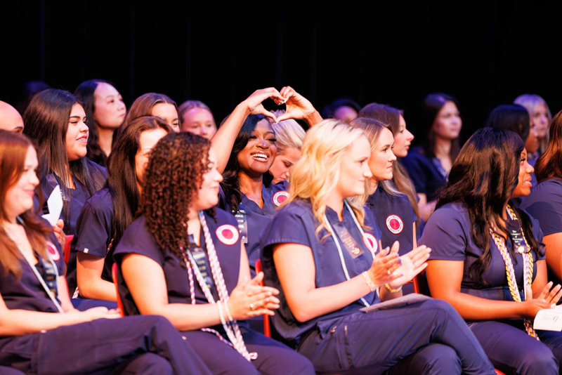 A large group of students in matching navy scrubs sits on a stage or auditorium seating area during a ceremony. Several students are clapping, and one student near the center raises both hands overhead to form a heart shape. The group wears name badges and honor cords, with a dark theater-style backdrop behind them.