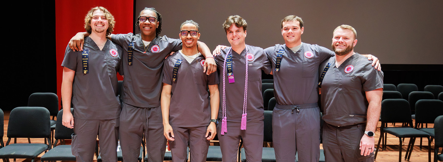 A group of nursing students in scrubs stand together on stage with arms around each other, posing for a celebratory photo.